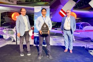 Individual holds a structural health monitoring award on stage at an international conference, with aircraft exhibits and event branding visible in the background.